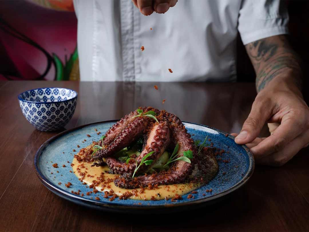 Chef garnishing grilled octopus on a blue plate.