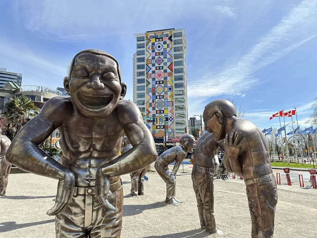 Bronze statues of laughing figures at A-Maze-Ing Laughter near English Bay, Vancouver, with colorful mural in the background.