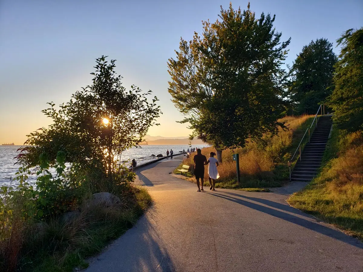 Couple walking along English Bay Beach at sunset, with warm light casting long shadows and ocean views in the background.